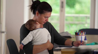 Anne bir masada otururken bebeğini kucağında tutuyor, bakıcılık ve iş arasında çoklu görev yapıyor, sakin bir ev ofisinde görevleri tamamlamaya odaklanıyor.