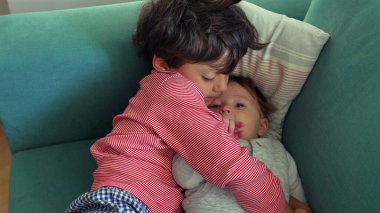 Ağabey, kanepede yan yana uzanırken bebeğin elini öpüyor, yakın kardeş sevgisi ve ilgi anı, bağlanma ve şefkat ile sıcak bir yuva kuruyor.