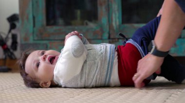 Anne bebeği giydirir, bebek yerde yatarken pantolon giyer, ellerin belini kapatır, bebek evde rutin bakım sırasında neşeyle gülümser.
