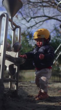 Küçük çocuk, kumları metal oyun yapısına aktarıyor ve merak koordinasyonunu, bağımsızlığını ve park ortamında duyusal öğrenimi ifade ediyor.