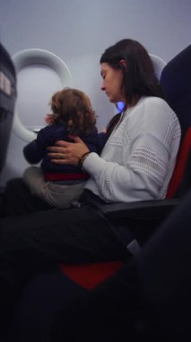 Ebeveyn, sabır denetimi ve yürümeye yeni başlayan bebekle seyahat etmenin günlük zorluklarını yansıtan küçük çocuğu uçak kabinine bağlar.