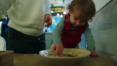 Genç çocuk yemek masasında tabağa uzanırken, anne de yanında durup güvenli keşif otonomisi büyümesini ve otantik aile etkileşimini destekliyor.