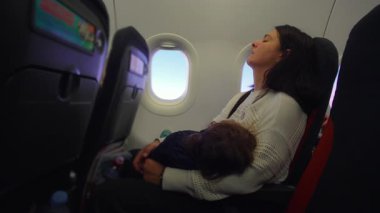 Anne uçuş sırasında dinleniyor. Çocuk kucağında uyuyor. Ebeveyn yorgunluğunu ifade ediyor. Şefkat gösteriyor.