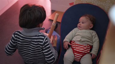 Bebeğimizin yanında otururken sandalyeyi ters tutan küçük çocuk çocuğa bakıyor, kardeş etkileşimine odaklanıyor ve kapalı mekândaki eğlenceli aile anlarına odaklanıyor.