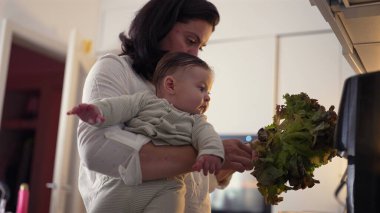 Anne mutfakta taze marul hazırlarken bebeği tutuyor, bebek dikkatle izliyor, aile etkileşimine ve yemek pişirme rutininde çoklu göreve odaklanıyor