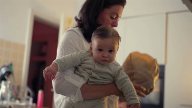 Anne kucağında bebek bakkaliyesi açarken, taze marul tutarken, bebek dikkatle izlerken, aile bağlarına ve mutfaktaki günlük ev aktivitelerine odaklanıyor.