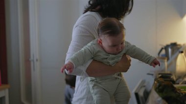 Anne mutfakta yemek hazırlarken bebeği kucağında tutuyor, bebek merakla etrafına bakıyor, genç çocuk başları eğik, mutfak sahnesi rutin bir şekilde yanında duruyor.