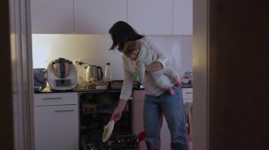 Anne mutfakta bulaşık yıkarken bebeği tutuyor, ev işleri sırasında birden fazla iş yapıyor, aile hayatına ve ebeveynlik sorumluluklarına odaklanıyor