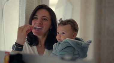 Anne kollarında bebekle dişlerini fırçalayıp aynaya bakıyor, sabah rutini, aile bağı, ilgili anne, banyo düzeneği, bebek gözlemleme, günlük temizlik uygulaması.