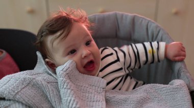 Yumuşak battaniyenin üzerinde eli ile gülümseyen bebeğe yakın çekim, neşeli ve neşeli yüz ifadesi, rahat bir şekilde oturup, çevreyle, yumuşak dokularla ve nazik anlarla meşgul olma