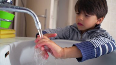 Çocuk banyo lavabosunda musluğun altında ellerini yıkıyor, hijyen ve temizliğe odaklanıyor, günlük kişisel bakım, sağlıklı alışkanlıklar öğreniyor, temiz ellerin yakın çekimi, su akışı