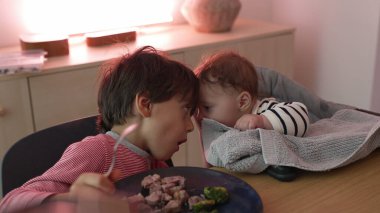 Bebeğe yakın olan genç bir çocuk, sevgi dolu bir jest, aile yemeği sırasında kardeş etkileşimi, hassas ve meşgul bir an, çocuklar arasında sıcaklık ve bağ.