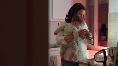 Anne, mutfak tezgahını düzenlerken bebeğini kucağında tutuyor, ebeveynliği ve ev işlerini dengeliyor, sıcak bir yuva ortamında bağ ve aile yaşamını geliştirmeye odaklanıyor
