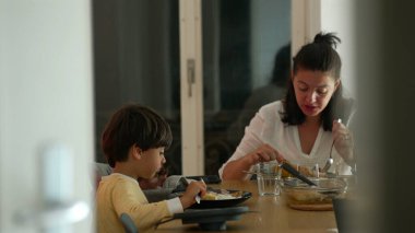 Anne ve küçük çocuk bir aile yemeğini paylaşıyor, yemek masasında doğal bir etkileşim, rahat bir atmosfer, aile bağlarını besliyor, ve evde günlük aile yaşamını paylaşıyor.