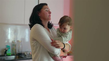 Anne mutfakta birden fazla iş yaparken bebeği kucağına alıyor, eşyalar düzenliyor ve ev sorumluluklarını yönetiyor, aile bakımı ve yetiştirme ortamlarına odaklanıyor