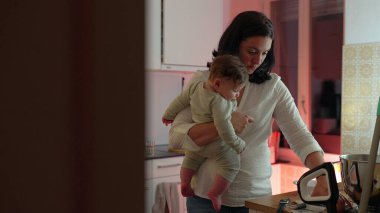 Anne, mutfak tezgahını düzenlerken bebeğini kucağında tutuyor, ebeveynliği ve ev işlerini dengeliyor, sıcak bir yuva ortamında bağ ve aile yaşamını geliştirmeye odaklanıyor