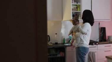 Anne, mutfak tezgahını düzenlerken bebeği kucağına alıyor, ebeveynlik ve ev işlerini dengelediğinde bağ kuruyor, sıcak ve ilgili bir ev ortamı yaratıyor.