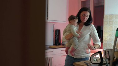 Anne, mutfak tezgahını düzenlerken bebeğini kucağında tutuyor, ebeveynliği ve ev işlerini dengeliyor, sıcak bir yuva ortamında bağ ve aile yaşamını geliştirmeye odaklanıyor