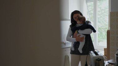 Anne mutfakta bebek beşiği yapıyor, sessiz bir bakım anında tezgahın yanında duruyor, ebeveynlik ve günlük ev işleri arasındaki dengeyi yansıtıyor.