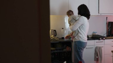 Anne, mutfak tezgahını düzenlerken bebeği kucağına alıyor, ebeveynlik ve ev işlerini dengelediğinde bağ kuruyor, sıcak ve ilgili bir ev ortamı yaratıyor.