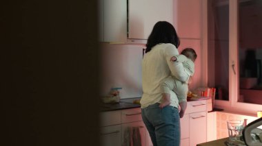 Anne, mutfakta taşınırken bebeğini kucağına alıyor, onu besliyor ve onunla ilgileniyor, günlük ev işlerini çocuğa ilgi ve şefkat ile dengeliyor.