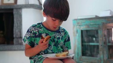 Genç bir çocuk tabağında peynirle ekmeği tutuyor, evde gündelik yemek saatlerinde özenle yiyor, sıcak bir aile ortamında yemeklerin tadını çıkarıyor ve beslenmeye odaklanıyor.