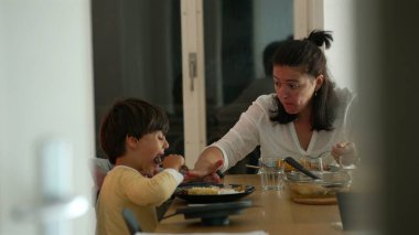 Anne ve küçük çocuk bir aile yemeğini paylaşıyor, yemek masasında doğal bir etkileşim, rahat bir atmosfer, aile bağlarını besliyor, ve evde günlük aile yaşamını paylaşıyor.