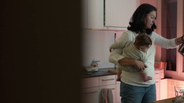 Anne, mutfakta taşınırken bebeğini kucağına alıyor, onu besliyor ve onunla ilgileniyor, günlük ev işlerini çocuğa ilgi ve şefkat ile dengeliyor.