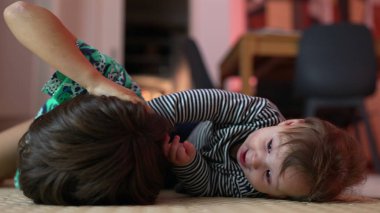 Bebek ve büyük kardeş yerde yatıyor, neşeli bir etkileşim içinde, bebek şakacı bir şekilde gülümserken kardeş saçı çekiyor, bağ kuruyor, aile bağlarını sıcak bir ortamda besliyor..