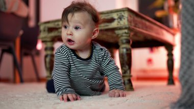 Bebek yerde yatıyor, elleri şaşırmış bir ifadeyle destekleniyor, kırsal ahşap masanın yanında mideye yakın vakit geçiriyor, çevreyi keşfediyor, erken gelişim motor aktiviteleri yapıyor.