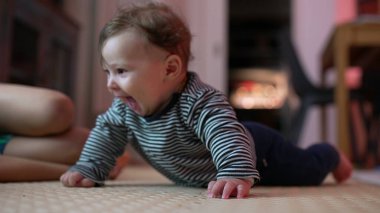 Bebek yerde yatıyor, kollarıyla yukarı itiyor, heyecanlı bir ifadeyle ağzı açık, neşeli bir an yaşıyor, erken motor becerilerini gösteriyor, kapalı alanda enerjik bir ortam.