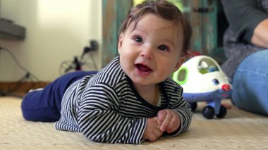 Bebek yerde neşeli bir ifadeyle yatıyor, kolları desteklenirken geniş bir şekilde gülümsüyor, mutluluk ve erken gelişim nişanı sergiliyor, arka planda görünen oyuncak uçak
