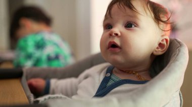 Bebek minderli bir sandalyede oturuyor, gözleri sonuna kadar açık, meraklı bir şekilde çevreyi keşfediyor, arka planda büyük kardeş, evde sessiz bir gözlemle meşgul.