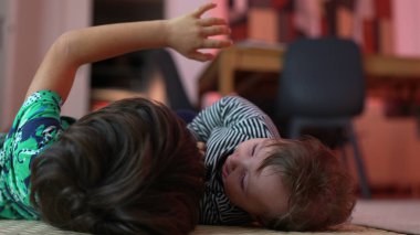 Bebek ve büyük kardeş yerde yatıyor, neşeli bir etkileşim içinde, bebek şakacı bir şekilde gülümserken kardeş saçı çekiyor, bağ kuruyor, aile bağlarını sıcak bir ortamda besliyor..