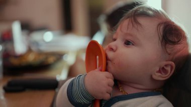 Bebek portakallı kaşık tutuyor, yanağa bastırıyor, gevşemiş ifade, yemek saatinde sandalyeye oturuyor, duyusal keşiflere ince odaklanıyor, neşeli bir evcil an.