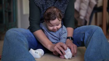 Bebek bakıcılığı anne kucağında, beyaz çoraplara odaklanmış nazikçe ayarlanmış, samimi bir bağ kurma anı sıcak aile ortamında bakım bakımını ön plana çıkarıyor.