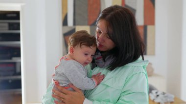 Anne kucağında neşeli bir gülümsemeyle bebeği tutarken bebek biraz sıkıntılı görünüyor, anne mutluluğu ile bebek duygusal ifadesi arasındaki zıtlığı yürekten bir aile anında yakalıyor.