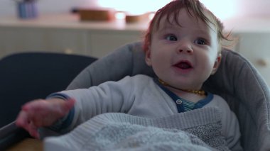 Bebek yüksek sandalyede oturuyor, parlak gözlerle gülümsüyor, neşeli ve çekici bir ifade, yumuşak bir battaniyeye sarılmış, sıcak bir aile ortamı, sıcaklık ve günlük hayatta mutluluk.