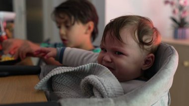 Bebek yüksek sandalyede oturuyor, gittikçe hayal kırıklığına uğruyor, kaşları çatılmış, şikayet etmeye başlıyor, rahatsızlık gösteriyor, büyük kardeş arka planda yemek yiyor, sabırsız bir an.