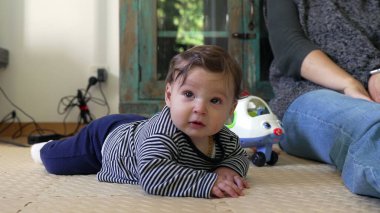 Bebek merakla yerde yatıyor, ön kollara dayanıyor, çevreyi gözlemliyor, oyuncak uçak yakınlarda, erken gelişim ve keşif gösteriyor.