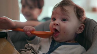 Bebek yüksek bir sandalyede otururken büyük turuncu bir kaşık tutuyor, merakla etrafı keşfediyor, büyük kardeş arka planda bulanıklaşıyor, aile yemeği zamanı ortamı, eğlenceli etkileşimler