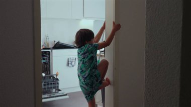 Genç bir çocuk kararlı bir ifadeyle kapı çerçevesine tırmandı, elleri ve ayaklarıyla kavrıyor, eğlenceli güç ve koordinasyon gösterisi, rahat mutfak ortamında maceracı bir iç mekan.