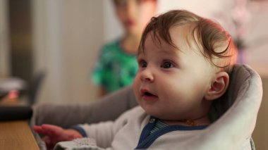 Bebek minderli bir sandalyede oturuyor, gözleri sonuna kadar açık, meraklı bir şekilde çevreyi keşfediyor, arka planda büyük kardeş, evde sessiz bir gözlemle meşgul.