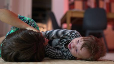 Bebek ve büyük kardeş yerde yatıyor, neşeli bir etkileşim içinde, bebek şakacı bir şekilde gülümserken kardeş saçı çekiyor, bağ kuruyor, aile bağlarını sıcak bir ortamda besliyor..