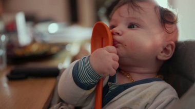 Bebek portakallı kaşık tutuyor, yanağa bastırıyor, gevşemiş ifade, yemek saatinde sandalyeye oturuyor, duyusal keşiflere ince odaklanıyor, neşeli bir evcil an.