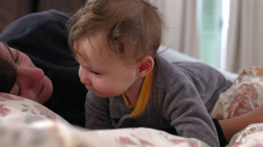 Bebek kameraya doğrudan meraklı bir ifadeyle bakarken anne yan yana uzanmış, meşgul ve dikkatli, samimi aile anı, kapalı alanda yakın bağ, erken çocuk gelişimi