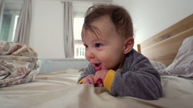 Bebek yatakta yüzüstü yatar, kameraya doğrudan meraklı ve meşgul bir ifadeyle bakar, erken motor beceri geliştirme, sıcak aile ortamı, keşif ve büyüme