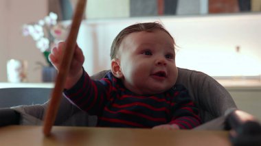 Bebek yüksek sandalyede oturuyor, bir testere tutuyor ve ısırmaya çalışıyor, odaklanmış ifade, keşfetme dokusu, erken duyusal deneyim, kapalı bir aile ortamı