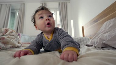 Bebek yatakta yüzüstü yatıyor, meraklı ve oyuncu bir ifadeyle ileri uzanıyor, erken motor beceri geliştirme, kapalı bir aile ortamı, erken çocuklukta keşif ve büyüme