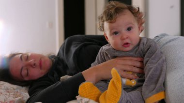 Bebek yatağında dinlenirken anne kolunda oturuyor, kameraya tuhaf bir ifadeyle bakıyor, rahat bir aile anı, evde huzurlu bir ortam, rahatlatıcı bir atmosfer.